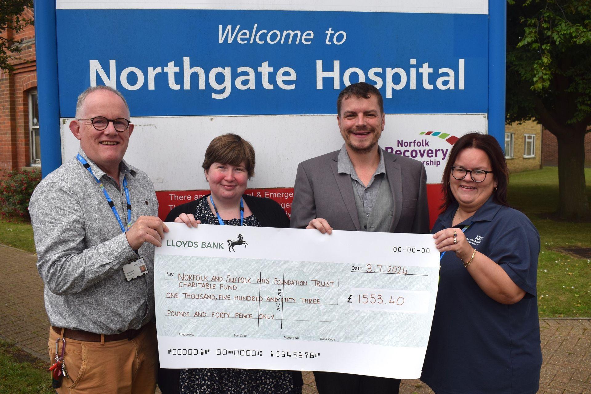 Four people holding a large donation check outside Northgate Hospital with a sign in the background.